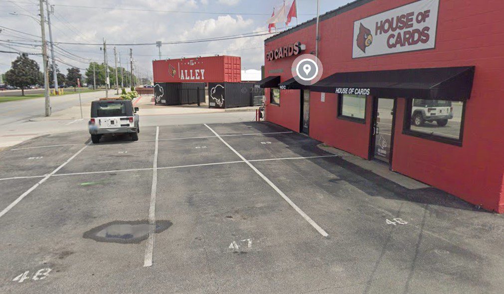 A red and black building labeled "House of Cards" sits next to an empty parking lot. In the background, a red structure with a cardinal logo and the word "Alley" is visible. A white vehicle is parked on the left.