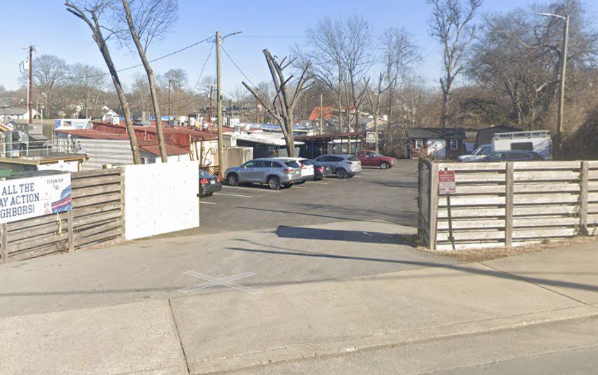 A small parking lot with several cars parked, surrounded by a wooden fence. Leafless trees and small buildings are visible in the background under a clear sky.