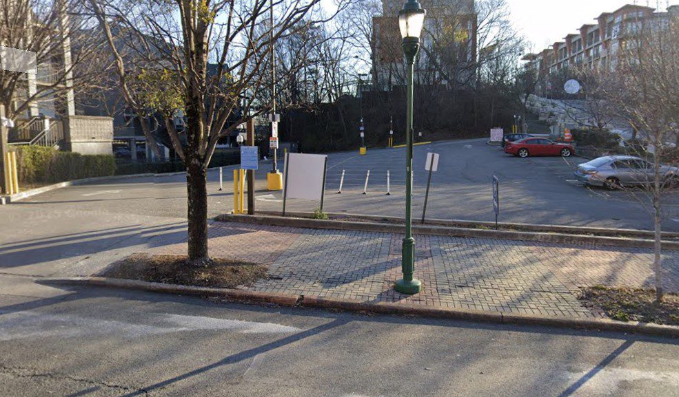 A mostly empty parking lot with a few scattered signs and parking barriers. Trees with bare branches line the area, and a lamppost stands near a brick-paved sidewalk in the foreground. Buildings are visible in the background.