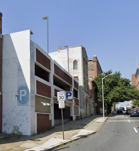 A partially dilapidated multi-story parking garage stands on a sloped city street with a speed limit sign reading "30 MPH." Trees line the sidewalk further down the street under a clear blue sky.