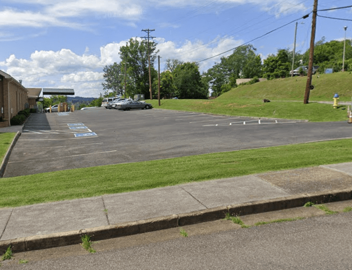 A mostly empty parking lot with several handicap parking spaces near a brick building, a few cars parked in the distance, green grass on both sides, and trees and utility poles in the background under a partly cloudy sky.