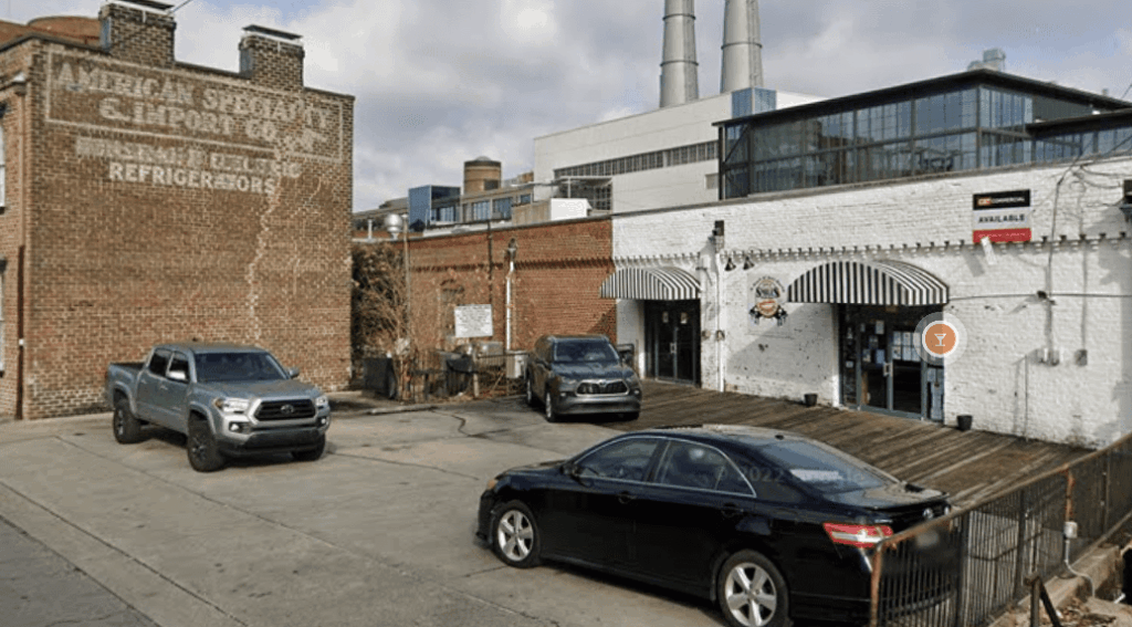 Three cars parked in a Parking Management Company parking lot beside brick buildings. One building has a faded painted sign, another features black and white striped awnings and an "available" sign. Factory smokestacks visible in the background.
