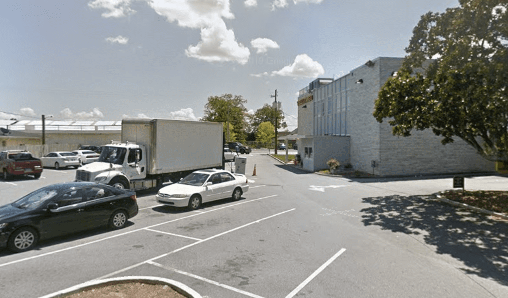 A small parking lot with several cars and a white box truck. A grey building with large windows is on the right, and trees and a blue sky with clouds are visible in the background.