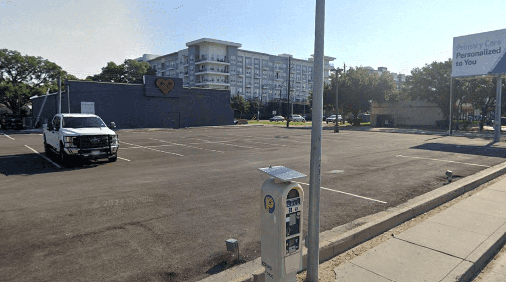 A mostly empty parking lot with a single white vehicle, a parking meter in the foreground, and a gray building with a heart mural. An apartment complex and trees are in the background.