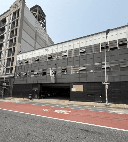 A three-story parking garage with a large 'P' sign above the entrance sits next to a tall building. The street in front has a red bus lane and white bike lane markings.