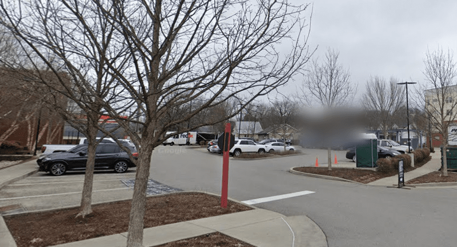 A parking lot on a cloudy day with several parked cars, leafless trees, orange traffic cones, and surrounding buildings in the background. A sidewalk runs along the edge of the lot.