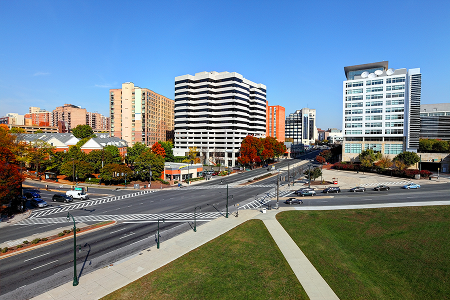 A wide city intersection with multiple lanes and crosswalks, bordered by modern high-rise buildings, trees with autumn foliage, and a clear blue sky overhead. Cars are stopped at the traffic lights.