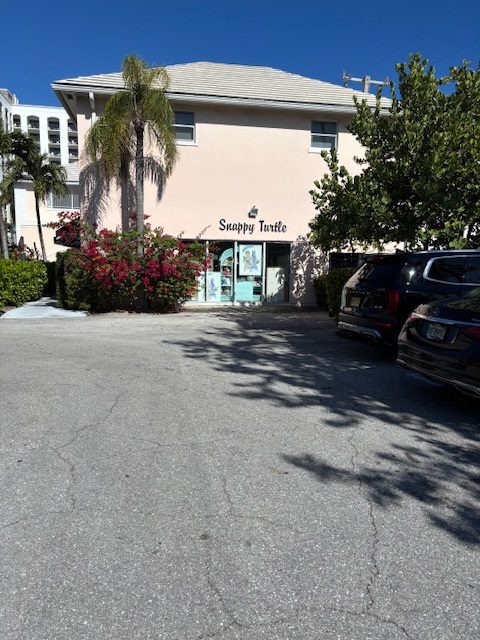 A pale pink building with a sign reading "Snappy Turtle" above aqua double doors, surrounded by green bushes, palm trees, and parked cars under a clear blue sky.