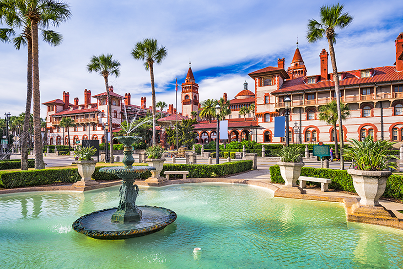 A large ornate fountain sits in a landscaped plaza with palm trees, in front of a grand historic building with red roofs and towers under a blue sky.