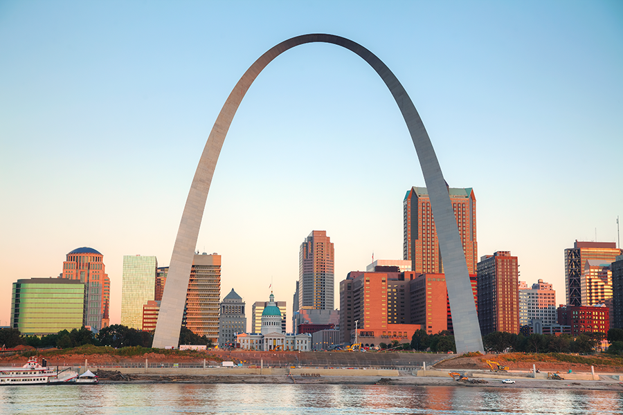 The Gateway Arch rises over the downtown St. Louis skyline at sunrise, with modern buildings and the Mississippi River in the foreground.