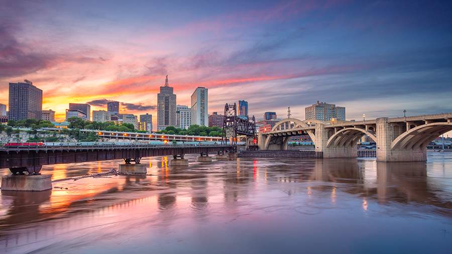 A city skyline at sunset with colorful clouds, tall buildings, and bridges spanning a calm river, reflecting the sky and structures in the water.