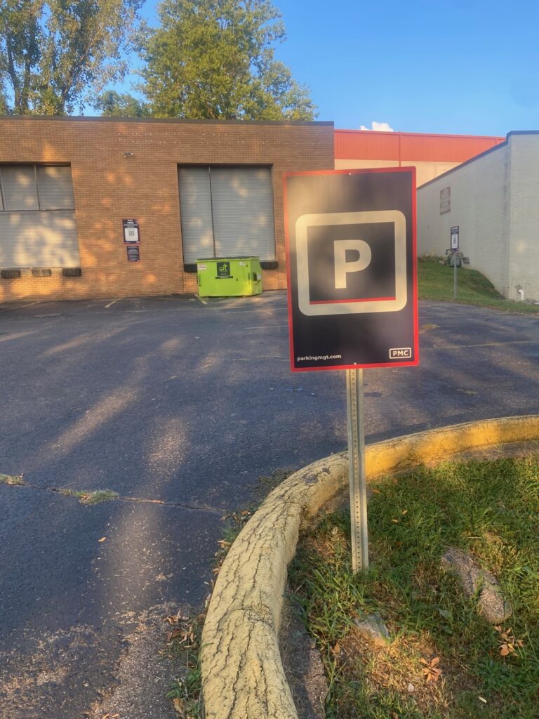 A black and white parking sign with a large "P" stands on a curb in front of a brick building and a green dumpster, with afternoon sunlight casting shadows.