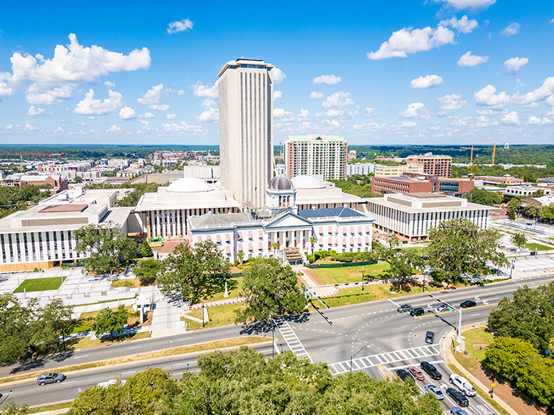 Aerial view of the Florida State Capitol complex in Tallahassee, featuring the historic capitol building with a dome, the modern capitol tower, and surrounding cityscape under a blue sky with scattered clouds.