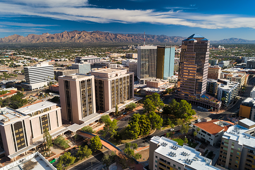 Aerial view of downtown Tucson, Arizona, with modern office buildings, tree-lined streets, and distant desert mountains under a blue sky with scattered white clouds.