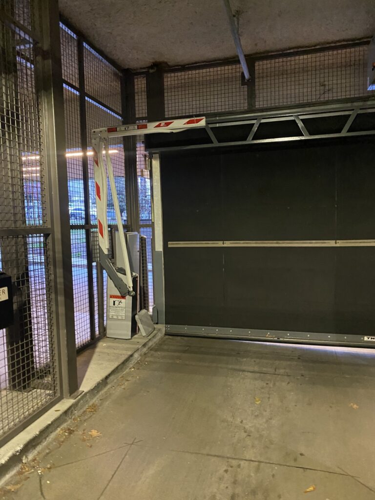 A metal security gate, partially open, blocks the entrance to a parking garage. The surrounding walls are made of metal mesh, allowing some light through. The ground is concrete with a few scattered leaves.