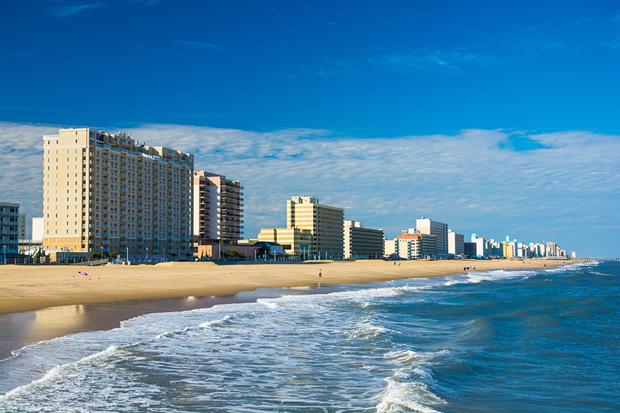 High-rise hotels and buildings line a sandy beach next to the ocean under a blue sky with scattered clouds, with gentle waves rolling onto the shore.