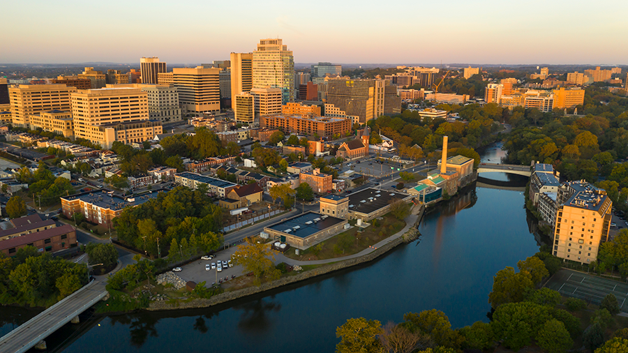 Aerial view of a city with tall buildings, a river running through the center, tree-lined streets, and a hazy sky at sunset.