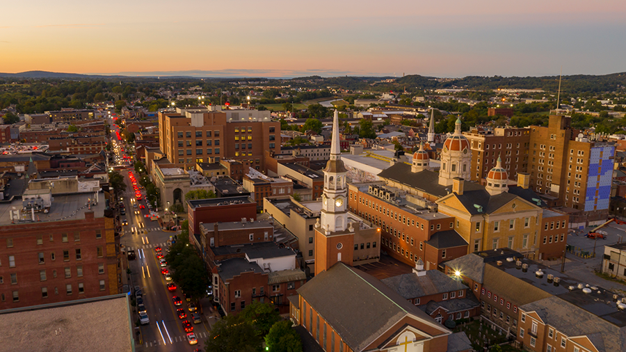 Aerial view of a city at sunset, featuring historic buildings, a church with a tall steeple, traffic on the streets, and green hills in the distant background.
