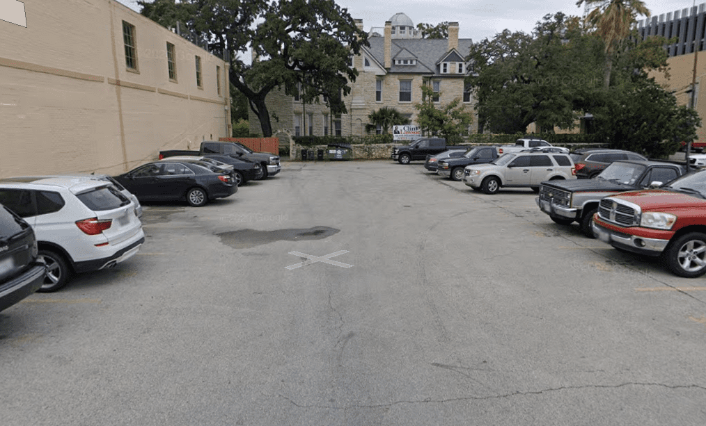 A small outdoor parking lot with cars parked on both sides, surrounded by trees and buildings, including a historic-looking stone house in the background.