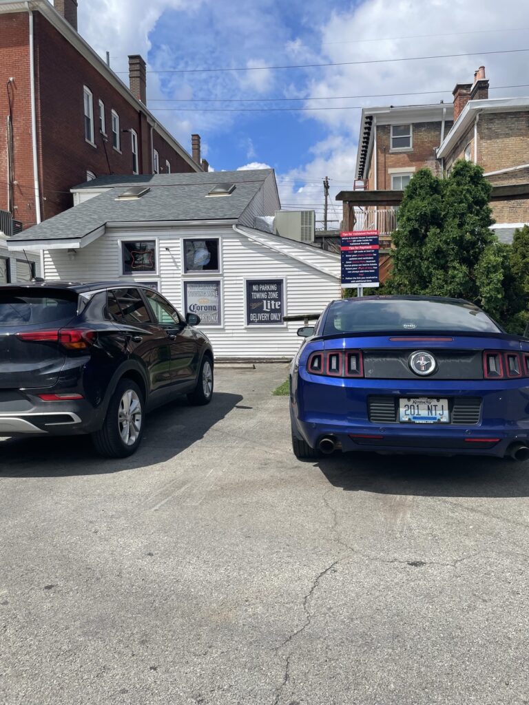 Two cars are parked in front of a small white building with signs in the windows. The building is surrounded by brick structures, and the sky is partly cloudy.