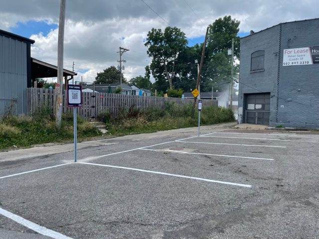 An empty parking lot with freshly painted white lines, two signs with QR codes, and a gray building on the right with a "For Lease" sign. Overgrown grass and wooden fence are visible in the background under a cloudy sky.