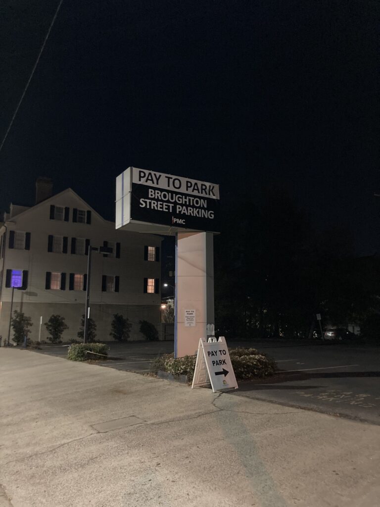 A Parking Management Company-owned parking lot at night with a large sign reading “PAY TO PARK BROUGHTON STREET PARKING.” A smaller board with the same message is placed near the entrance. A building with lit windows is in the background.