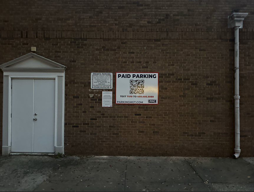 A brick wall with a white door on the left and three PMC signs, including a large "Paid Parking" sign with a QR code and instructions to text a number to pay for parking.