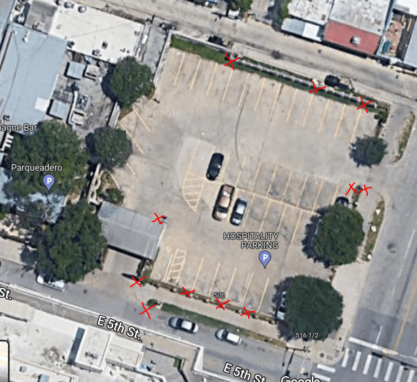 Aerial view of a mostly empty parking lot labeled “HOSPITALITY PARKING,” with a few cars parked and red X marks on many parking spaces and surrounding spots. Trees line parts of the lot and nearby streets.