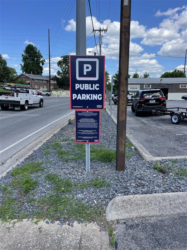 A sign labeled "Public Parking" stands beside a street, near a parking lot with several vehicles. The sky is blue with scattered clouds, and there are buildings and power lines in the background.