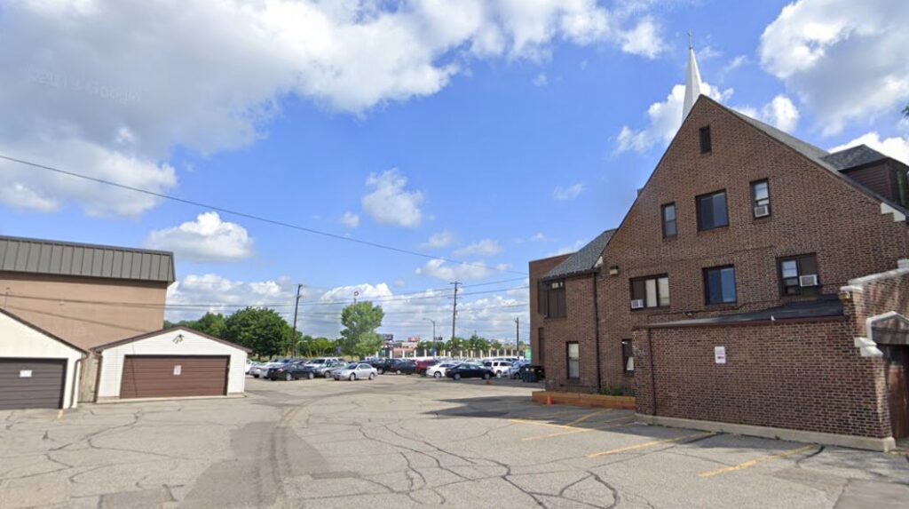 PMC parking lot with several cars, bordered by a brown brick building on the right and two small garages on the left.