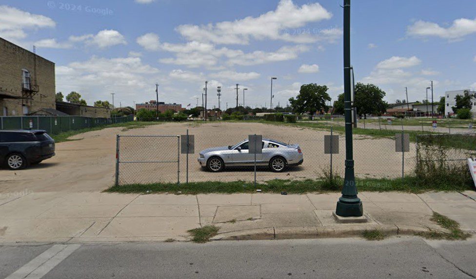 A fenced-in PMC parking lot with one silver convertible and another black car on the left. A green streetlamp and sidewalk are in the foreground; buildings and trees are visible in the background under a partly cloudy sky.