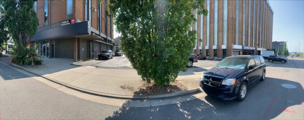 A panoramic street view of a commercial building with a tree in the center foreground, cars parked along the sidewalk, and clear blue sky in the background.