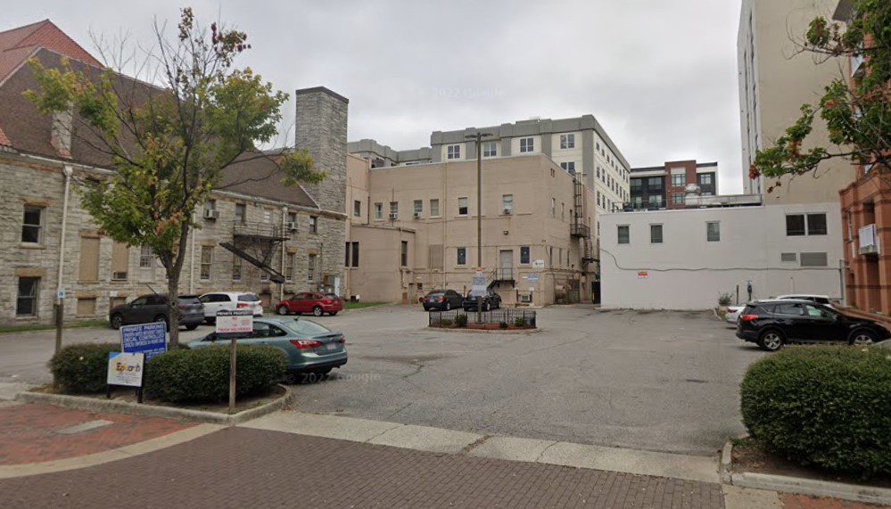 A PMC-owned urban parking lot with a few cars parked. Surrounding the lot are older stone and brick buildings, and some bushes line the edges. The sky is overcast.