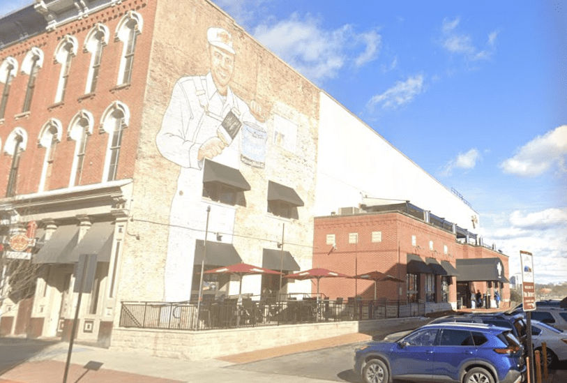 A brick building features a large mural of a smiling man in a uniform holding a bucket. Below, a restaurant patio with red umbrellas and several parked cars line the street under a blue sky with scattered clouds.