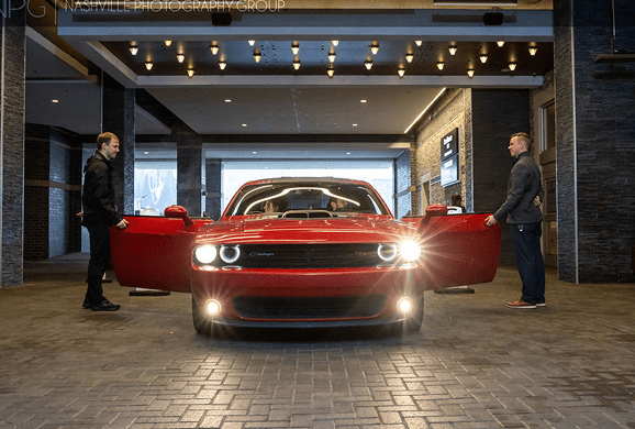 A red Dodge Challenger with headlights on is parked under a covered entryway. Two men stand on either side, each holding a door open. The background features brick walls and ceiling lights.