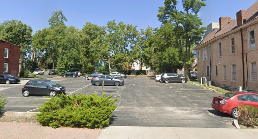 A nearly empty parking lot with a few cars parked, surrounded by trees and older brick buildings on a sunny day. Shrubs and a sidewalk are visible in the foreground.