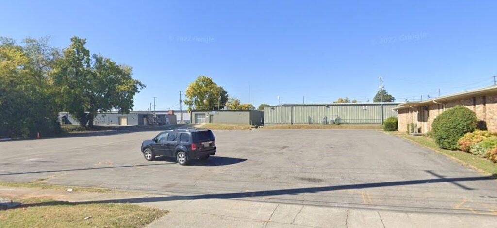 A mostly empty parking lot with a single black SUV parked near the center. There are industrial buildings and greenery in the background under a clear blue sky.