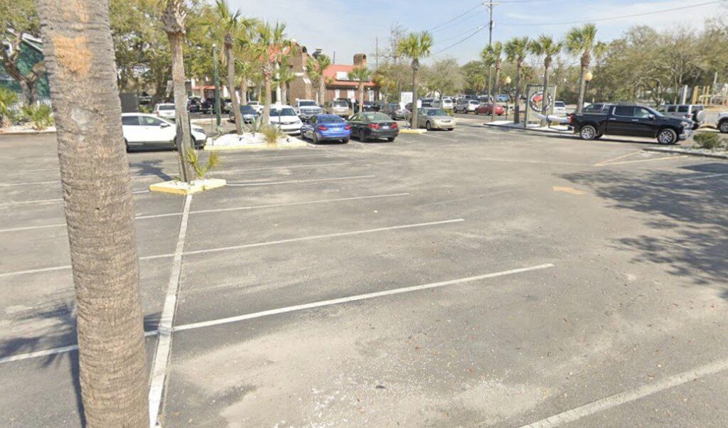 A nearly empty parking lot with a few cars parked near the back. Palm trees line the lot and some buildings and trees are visible in the background under a partly cloudy sky.