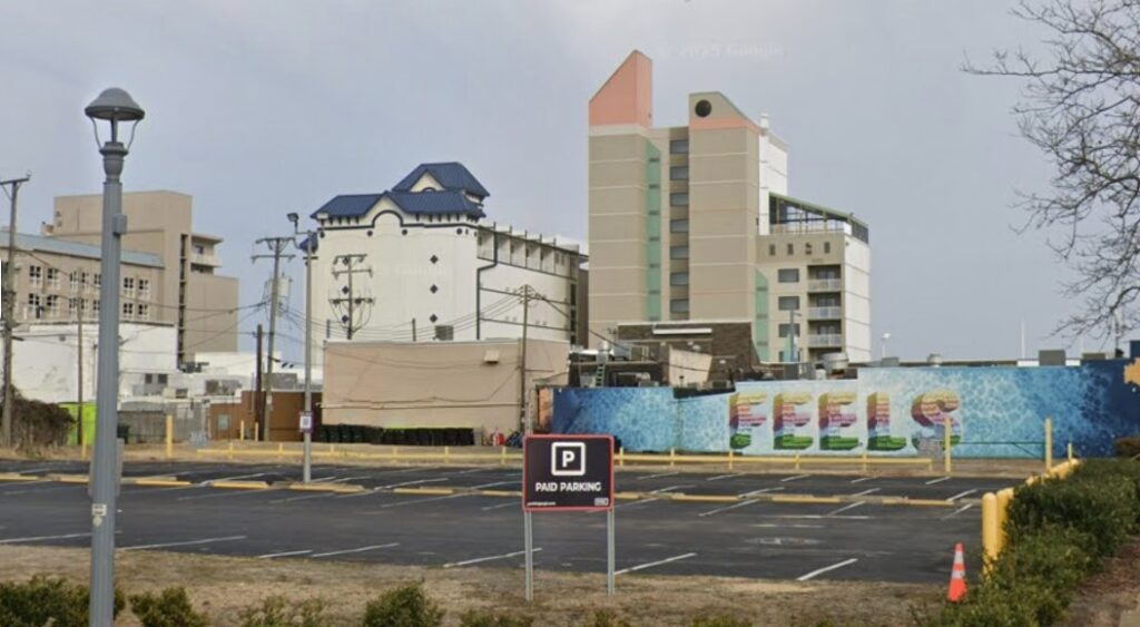 A mostly empty paid parking lot with a PMC parking sign in front, bordered by a blue mural and several tall buildings in the background.