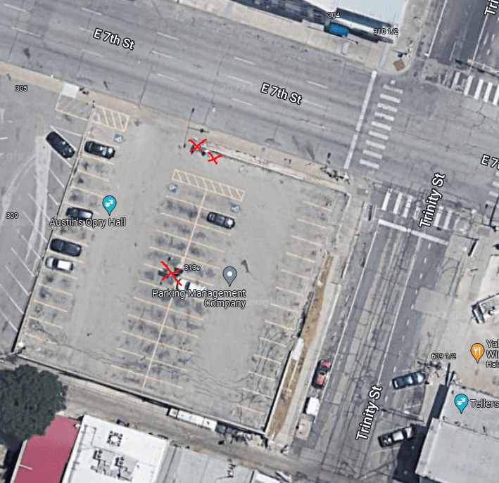 Aerial view of a parking lot at the corner of E 7th St and Trinity St, with several parked cars and three red X marks on empty parking spaces near the center and top left side of the lot.