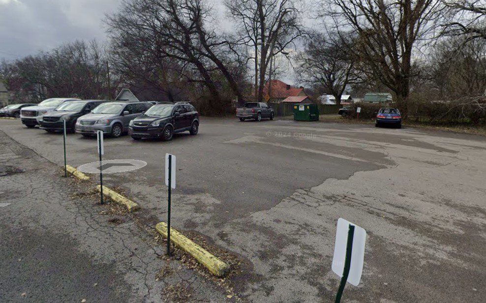 A mostly empty parking lot on a cloudy day, with six cars parked along the edge near trees and a green dumpster; yellow parking barriers and signposts are in the foreground.