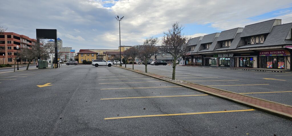 A mostly empty parking lot with a few cars parked near a row of shops on the right; small trees line the lot and the sky is partly cloudy.