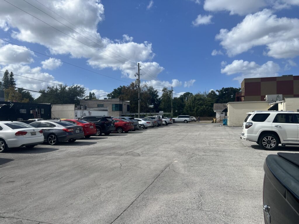 A parking lot with several parked cars on both sides, surrounded by trees and buildings under a partly cloudy blue sky.