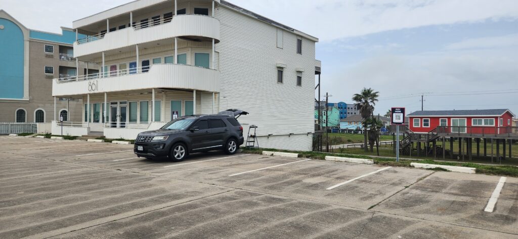 A gray SUV with its trunk open is parked in an empty lot near a white, three-story beach house. Colorful houses and palm trees are visible in the background under a partly cloudy sky.