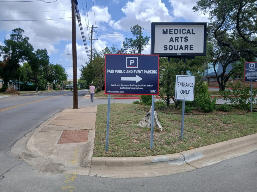 A sidewalk and street beside a parking lot with signs reading "Paid Public and Event Parking" and "Entrance Only" near a large "Medical Arts Square" sign; a person is walking away in the distance.