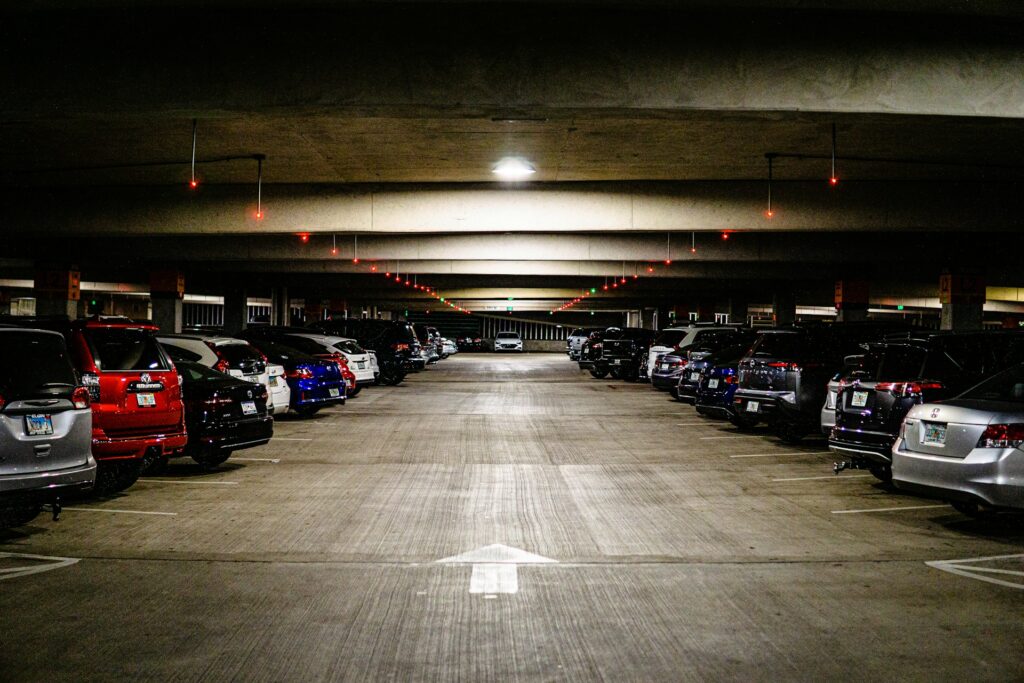 A well-lit underground parking garage owned by PMC with rows of parked cars on both sides, a concrete ceiling above, and red lights lining the ceiling. An arrow is painted on the concrete floor, pointing straight ahead.