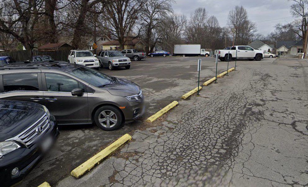 A cracked asphalt parking lot with several parked cars, yellow parking barriers, bare trees, and houses in the background under a cloudy sky.