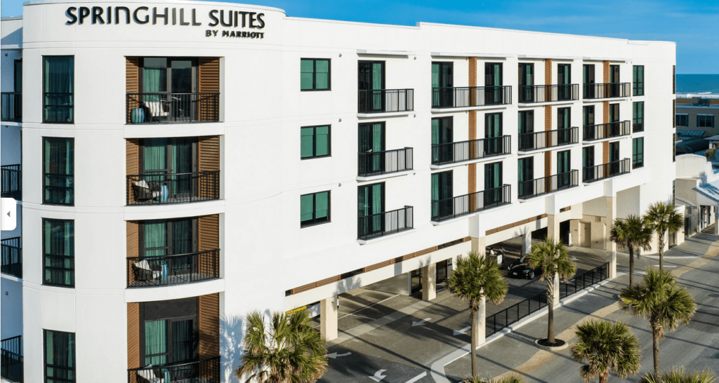 A modern, white four-story hotel labeled "SpringHill Suites by Marriott," with multiple balconies and large windows, palm trees in front, and a clear blue sky in the background.