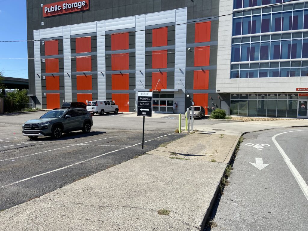 A nearly empty parking lot in front of a Public Storage building with red doors. Two vehicles are parked, and a sign in the lot reads "Public Parking." A bike lane runs along the edge of the lot.