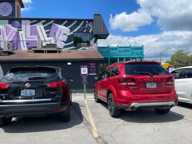 Two cars, one black and one red, are parked side by side in a parking lot. Behind them, a building features a colorful mural with purple and green designs and some visible text. The sky is partly cloudy.